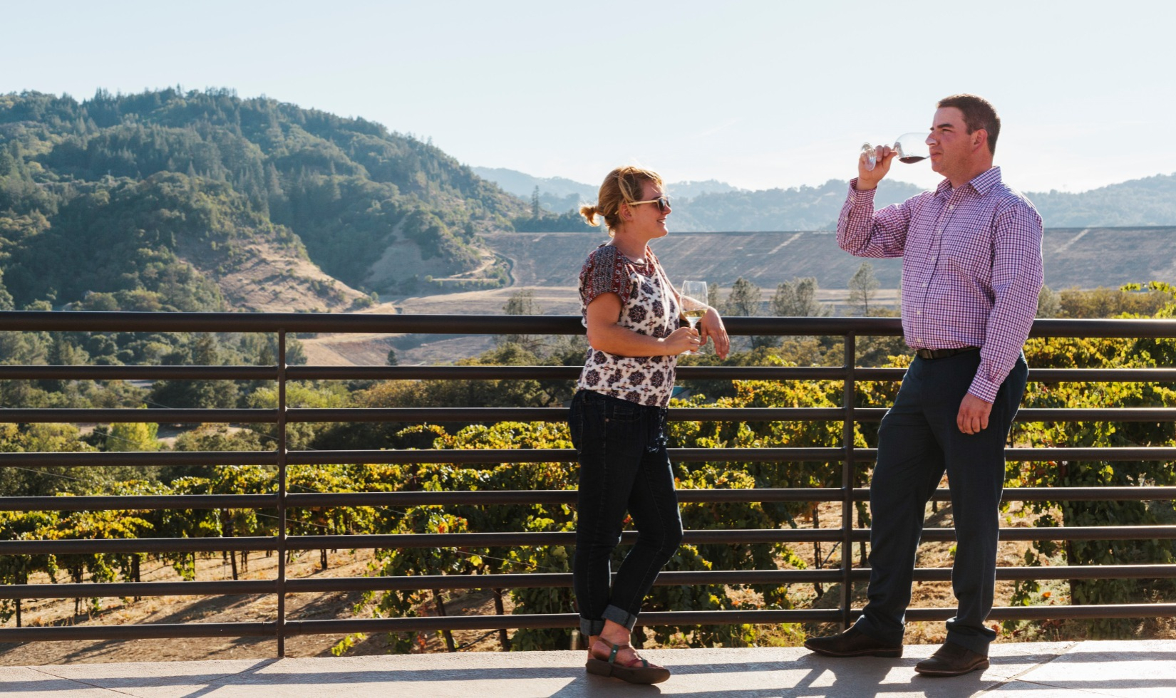 Couple drinking wine and standing by railing of terrace overlooking view of Dry Creek Valley vineyards