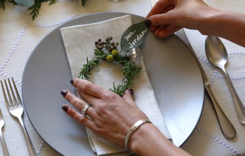 hands adjusting a rosemary wreath with a leaf name tag at a holiday table setting