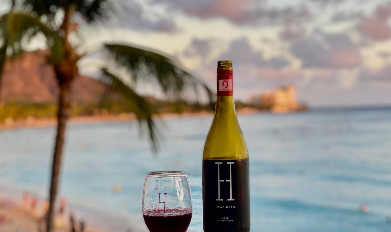 Bottle of Head High Pinot Noir with a stemless glass with wine on a balcony railing overlooking the beach and ocean in Hawaii. 