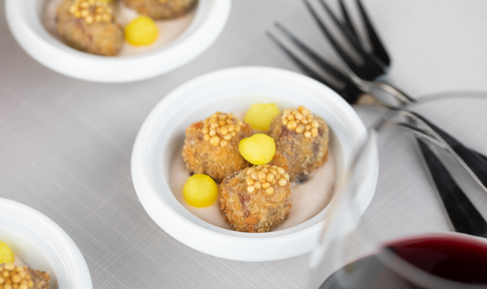small short rib bites with mustard seed on top in three white bowls paired with a glass of red wine and forks on dinner table