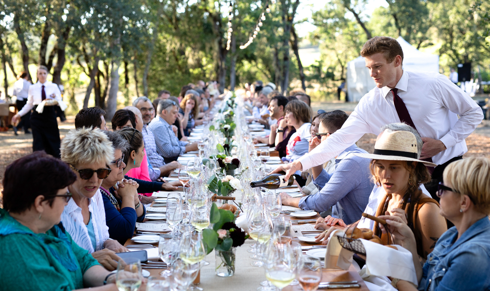 long dinner table with food and wine being poured in the middle of a vineyard