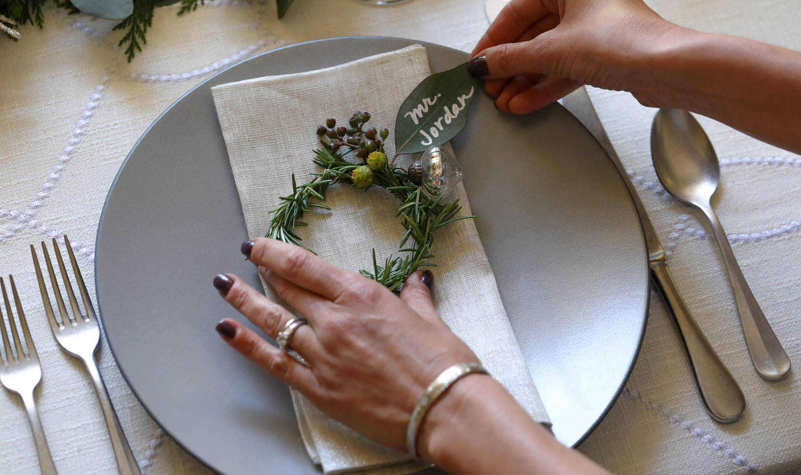 hands adjusting a mini rosemary wreath with a leaf name tag on a holiday place setting