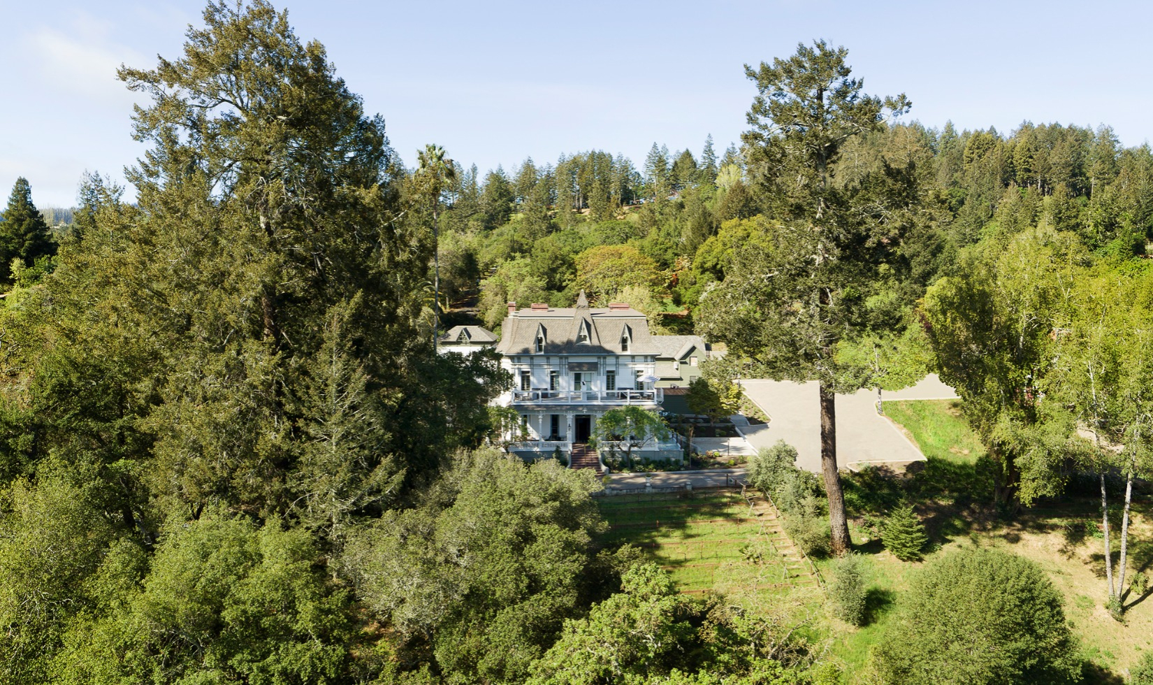 Aerial drone image of white Victorian mansion surrounded by green yards and forest.
