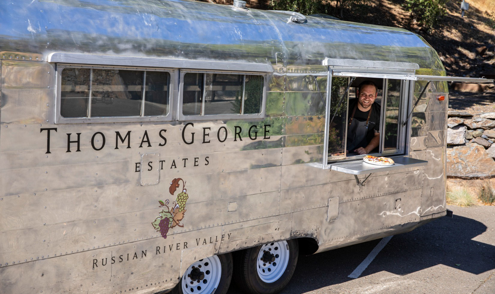 A silver airstream with the thomas george logo with a window open serving pizza