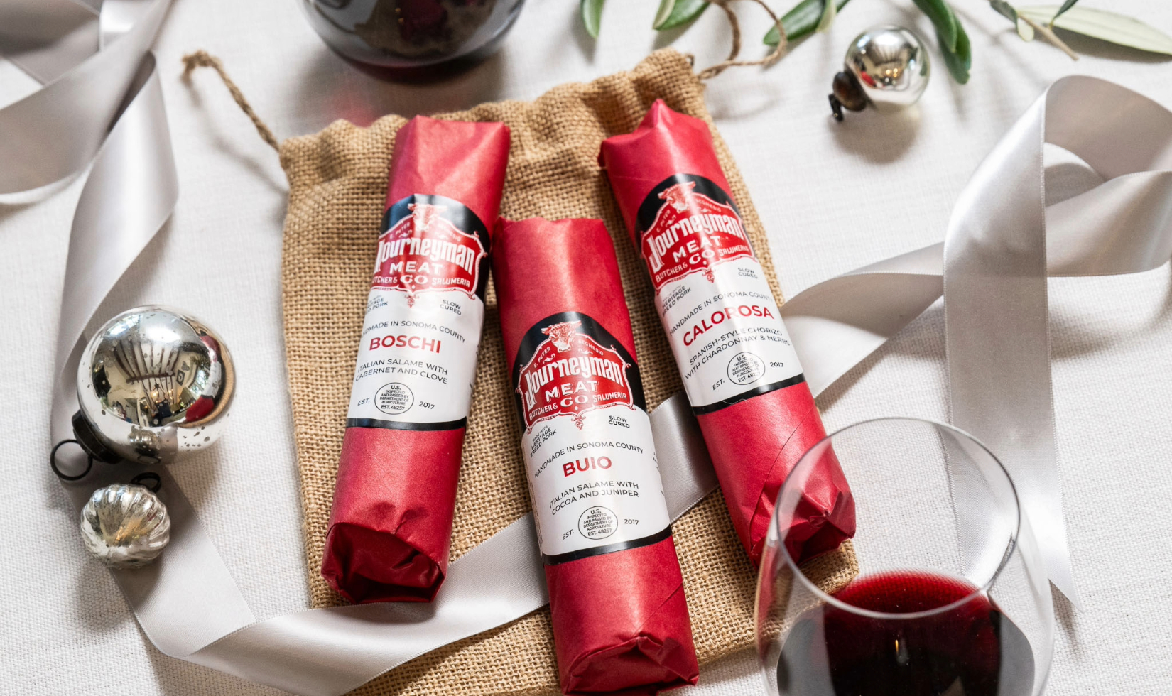 Three rolls of salumi wrapped in red packaging on table decorated with silver christmas ornaments and ribbon