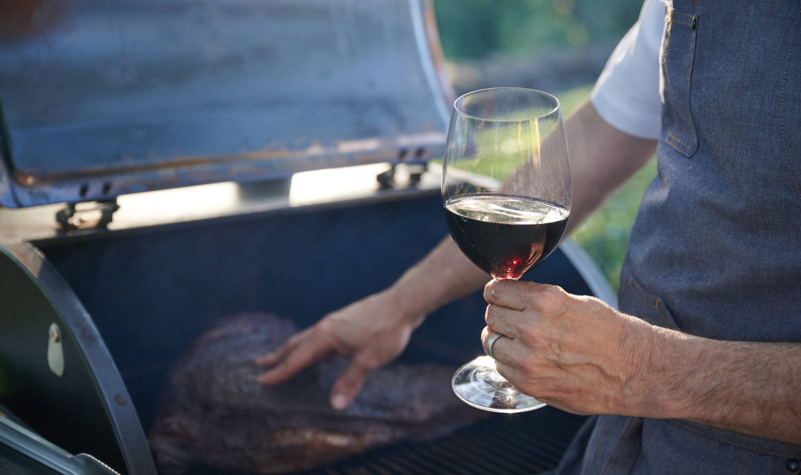 Hand touching meat on a grill with a glass of red wine in the other hand