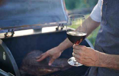 Hand touching meat on a grill with a glass of red wine in the other hand