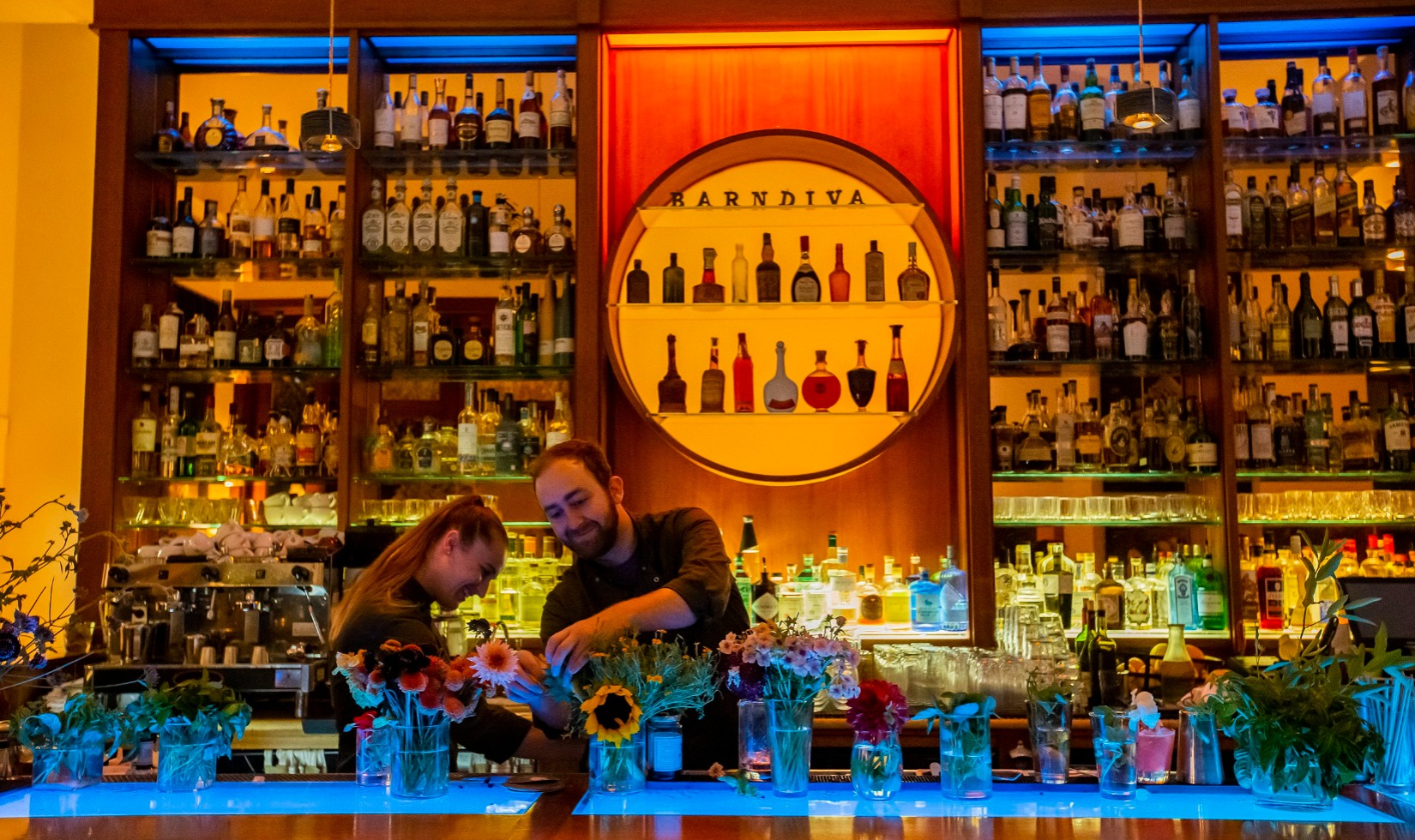 Bartenders mixing drinks in front of bar back-lit by blue lights with shelves lined with spirits bottles