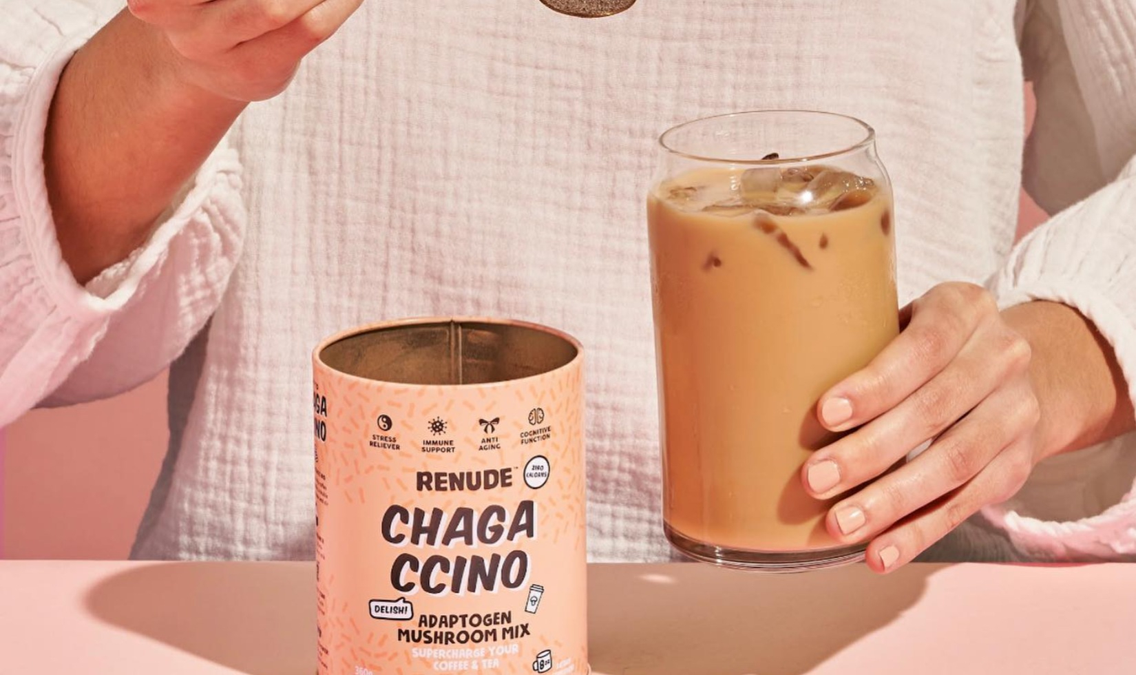 Cannister of Chagaccino next to glass of iced coffee held by a woman. 