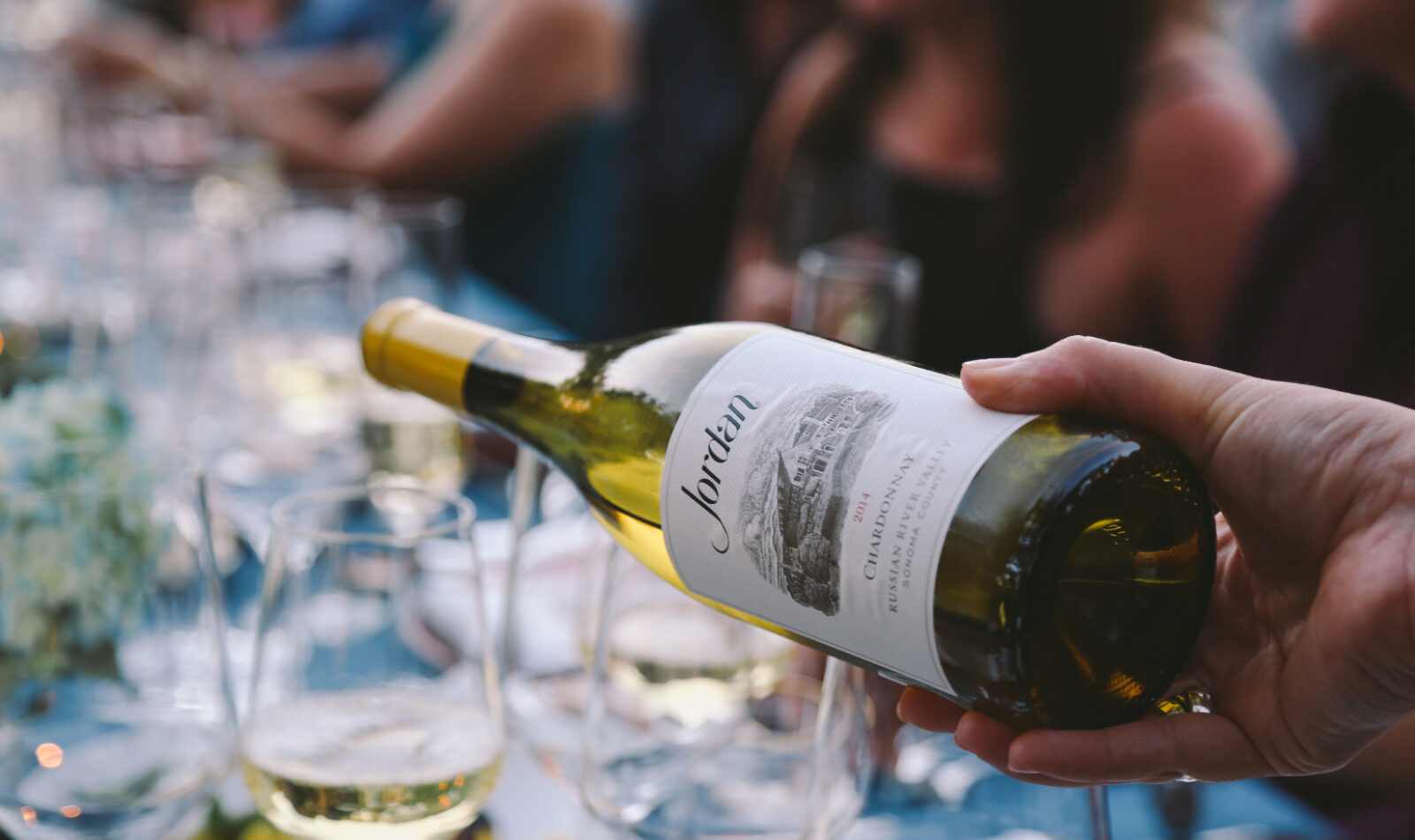 a person pouring a white wine to a glass in a table full of guests as featured in wine country table.