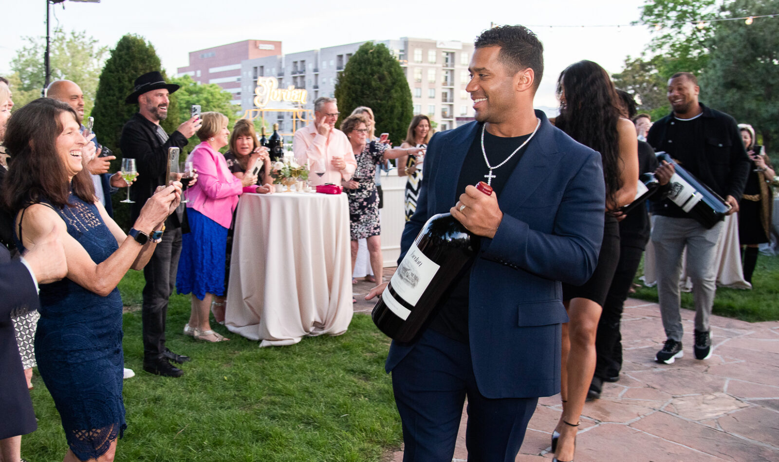 Russell Wilson celebrity walking through an outdoor party carrying a large bottle of wine with woman taking photos of him