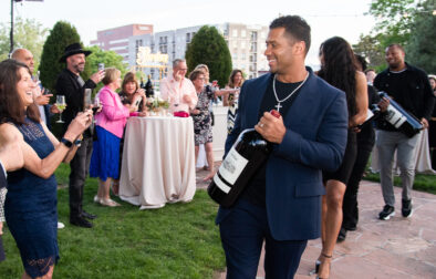 Russell Wilson celebrity walking through an outdoor party carrying a large bottle of wine with woman taking photos of him