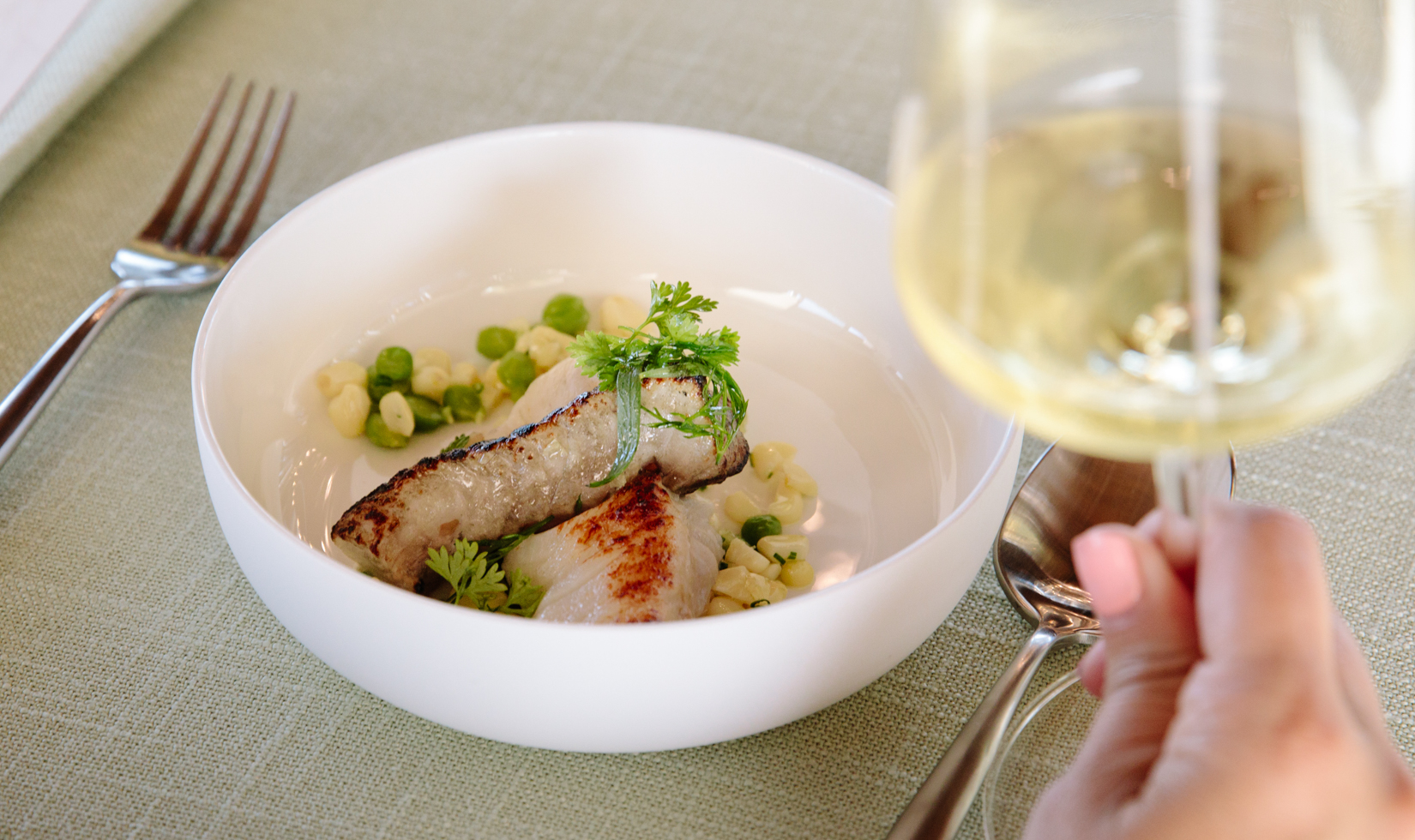 close up of halibut dish with a hand holding a glass of chardonnay wine 