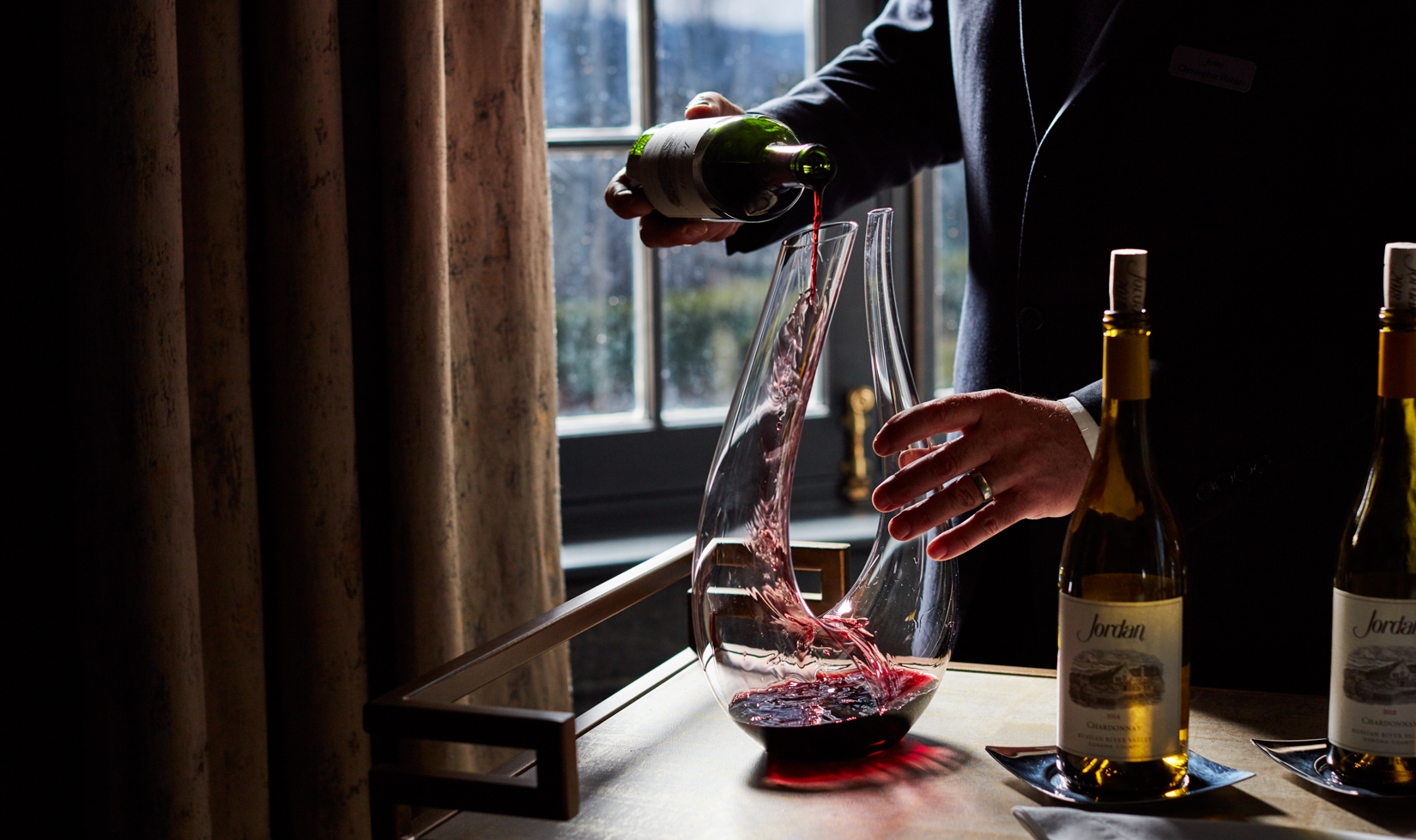 wine sommelier pouring red wine into glass decanter with window in background 