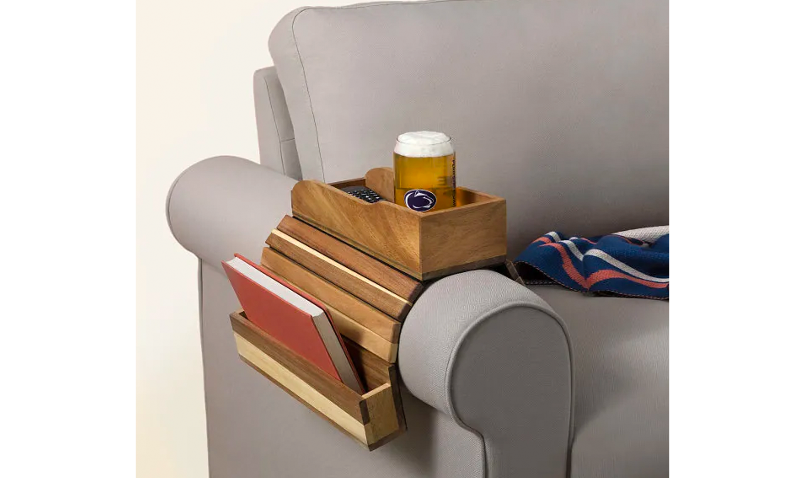 Wooden armrest organizer on a gray couch, holding a book, remote, and a glass of beer.