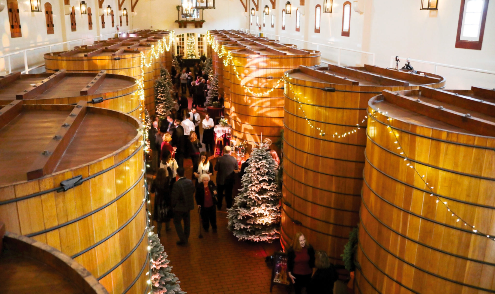guests walking through oak tank room at winery during christmas party with festive lights and christmas trees and garland